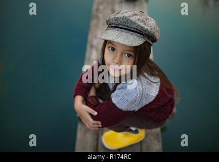 Portrait of child girl in cap and yellow boots at wooden bridge on background of river. Top view. Stock Photo