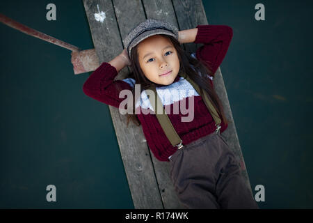 Child girl in cap lies on wooden bridge and smiles on background of river. Top view. Closeup. Stock Photo