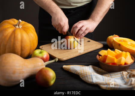 The cook cuts the apple into pieces for baking. On a wooden black table lie pumpkins of various sizes and shapes, ripe apples, a dishcloth and a bowl  Stock Photo