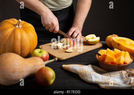 The cook cuts the apple into pieces for baking. On a wooden black table lie pumpkins of various sizes and shapes, ripe apples, a dishcloth and a bowl  Stock Photo