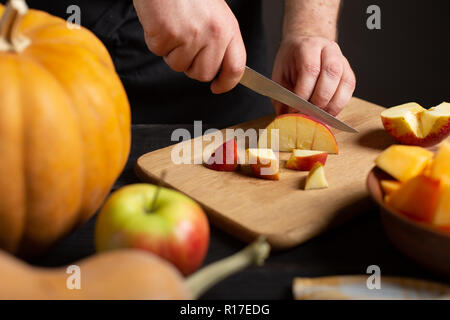 The cook cuts the apple into pieces for baking. On a wooden black table lie pumpkins of various sizes and shapes, ripe apples, a dishcloth and a bowl  Stock Photo