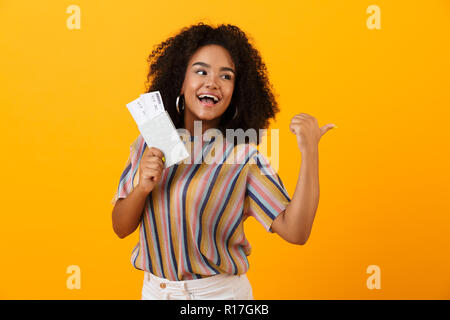 Portrait of a cheerful african woman standing isolated over yellow showing flight tickets with passport, pointing away Stock Photo