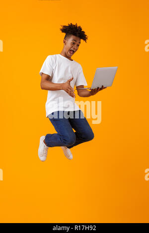 Image of happy excited young african man jumping isolated over yellow background using laptop computer. Stock Photo