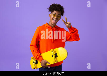Portrait of a cheerful young african man holding a skateboard isolated over violet background, showing peace Stock Photo