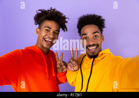 Two excited african man friends dressed in colorful hoodies standing isolated over violet background, taking a selfie Stock Photo