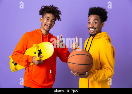 Two excited african man friends dressed in colorful hoodies standing isolated over violet background, holding skateboard and basketball, showing thumb Stock Photo