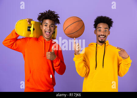 Two cheerful african man friends dressed in colorful hoodies standing isolated over violet background, holding skateboard and basketball, showing thum Stock Photo