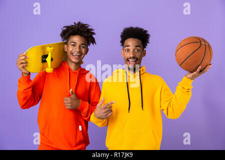 Two smiling african man friends dressed in colorful hoodies standing isolated over violet background, holding skateboard and basketball, showing thumb Stock Photo