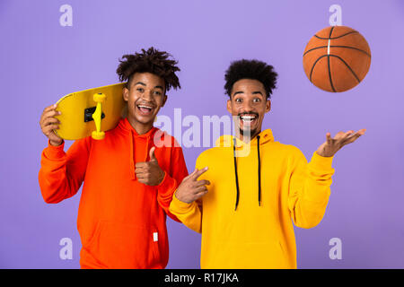 Two joyful african man friends dressed in colorful hoodies standing isolated over violet background, holding skateboard and basketball, showing thumbs Stock Photo