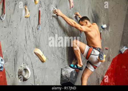 Male climber exercising in gym, climbing up on artificial wall with boulders. Stock Photo