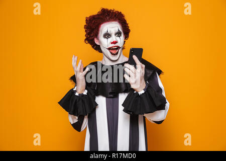 Surprised creepy man clown using smartphone isolated over orange Stock Photo