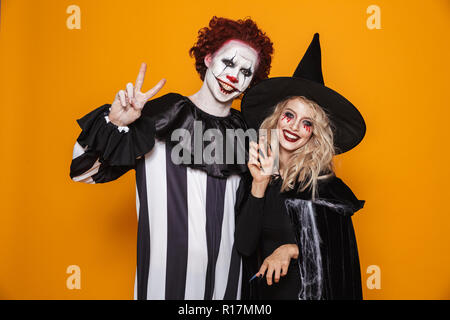 Cheerful woman witch and clown with hallowen make-up looking camera and smiling isolated over orange Stock Photo