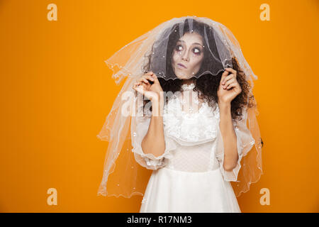 Thoughtful zombie bride in wedding dress and with halloween make-up looking up and holding veil isolated over orange Stock Photo