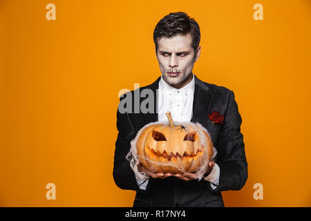 Displeased zombie man with halloween make-up and in suit holding pumpkin and looking aside isolated over orange Stock Photo