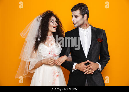 Cheerful dead bride and groom with halloween make-up looking at each other and smiling isolated over orange Stock Photo