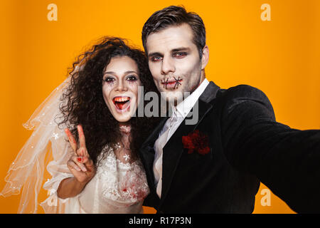 Funny pretty zombie bride and groom in halloween costumes making selfie at camera and smiling isolated over orange Stock Photo
