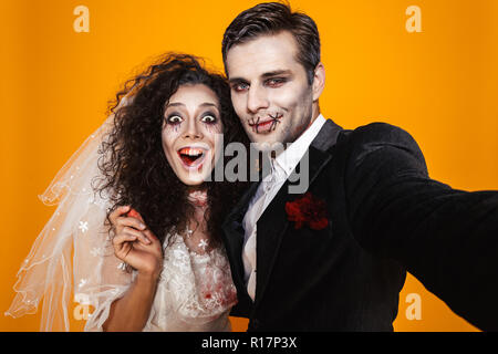 Photo of amusing zombie couple bridegroom and bride wearing wedding outfit and halloween makeup laughing while taking selfie isolated over yellow back Stock Photo