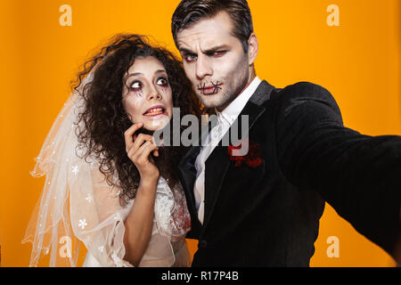 Scary funny bride and groom with halloween make-up making selfie at camera isolated over orange Stock Photo