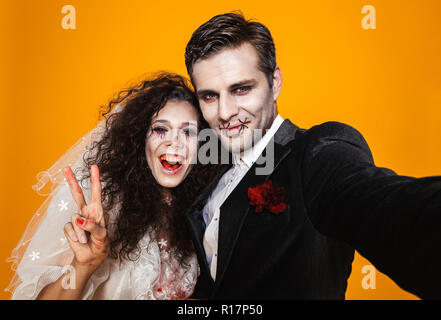 Photo of beautiful zombie couple bridegroom and bride wearing wedding outfit and halloween makeup laughing while taking selfie isolated over yellow ba Stock Photo