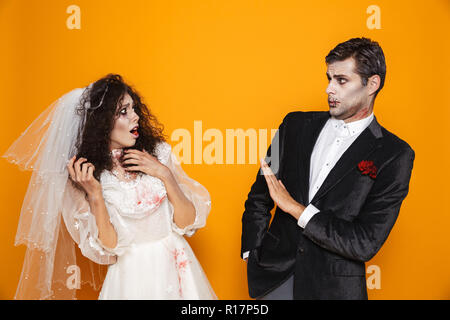 Surprised and scary bride and groom with halloween make-up looking at each other isolated over orange Stock Photo