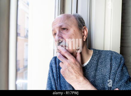 Old senior man looking throughout the window feeling confused and depressed alone at home in Aging Retirement Widower Dementia and Alzheimer concept. Stock Photo