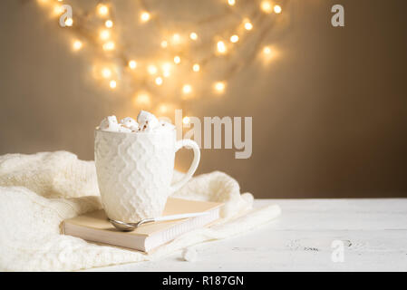 Hot chocolate with marshmallows in white mug,  copy space. Hot cocoa drink for Christmas and winter holidays with warm scarf, lights on white table. Stock Photo
