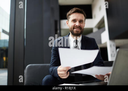 Happy bearded businessman with papers looking at you while working in modern office Stock Photo