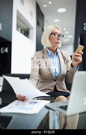 Busy elegant female agent sending message by smartphone while working with papers Stock Photo