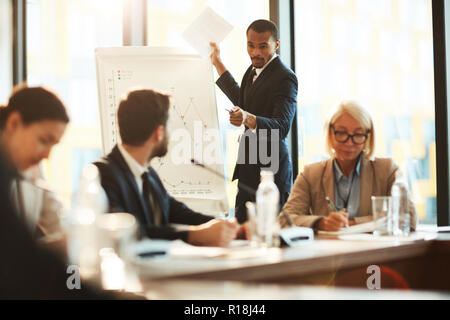 Confident African-american coach or reporter standing by whiteboard and pointing at one of attendants Stock Photo