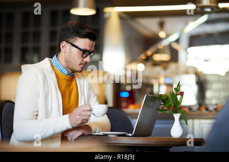 Young serious businessman reading data in laptop while having cup of tea or coffee in cafe Stock Photo