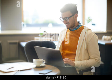 Happy young blogger with laptop sitting in cafe and browsing in the net by cup of coffee Stock Photo