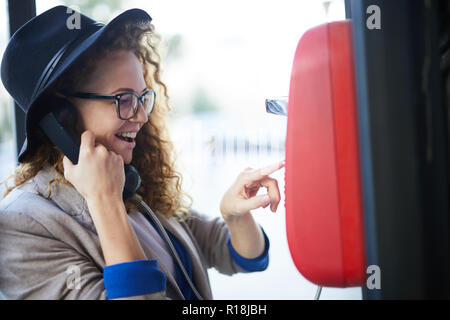 Young stylish female holding phone receiver by her ear while dialing number on panel of payphone in booth Stock Photo