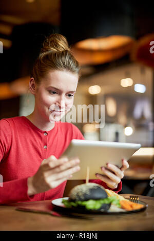 Pretty girl with touchpad sitting by served table in cafe and watching movie or video in the net Stock Photo