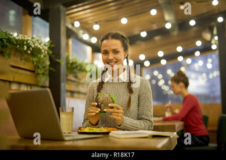 Smiling student with hamburger siting by table in front of laptop, having lunch and preparing for seminar in cafe Stock Photo