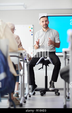 Young teacher in casualwear siting on chair in front of audience and introducing new topic to students Stock Photo