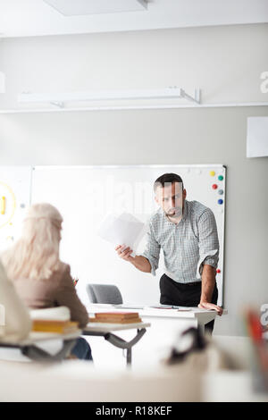 Serious or thoughtful teacher with papers leaning over his workplace in front of audience Stock Photo