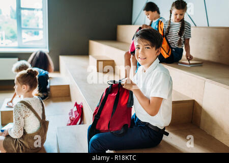 group of schoolchildren with backpack sitting on wooden tribune at school corridor Stock Photo