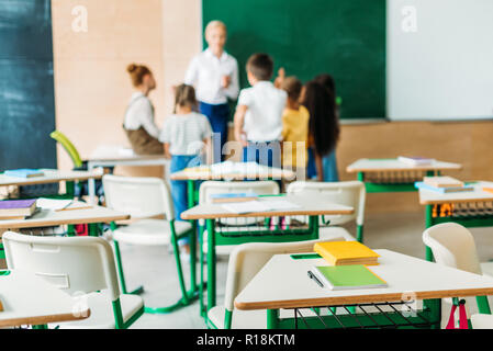 schoolchildren standing around teacher at classroom with desks on foreground Stock Photo