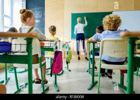 rear view of schoolchildren looking at teacher while she writing on chalkboard Stock Photo
