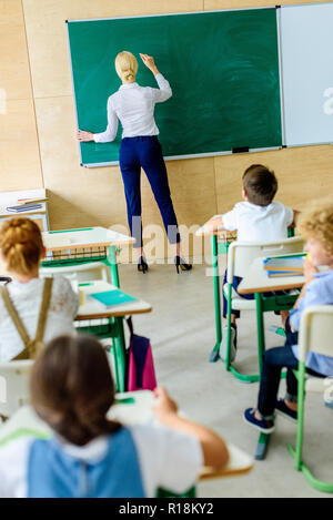 rear view of kids looking at teacher while she writing on chalkboard Stock Photo
