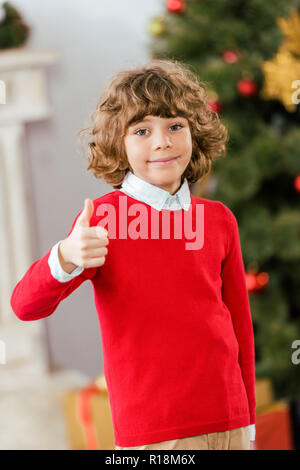 adorable happy kid showing thumb up with blurred christmas tree on background Stock Photo