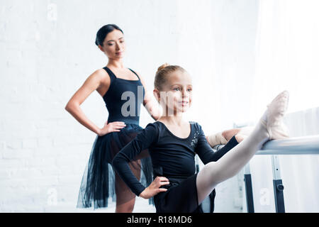 smiling little ballerina stretching while teacher standing behind in ballet school