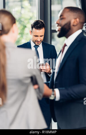 selective focus of businessman using smartphone while multiethnic colleagues talking in office Stock Photo