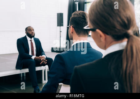 selective focus of african american speaker sitting on stage during seminar in conference hall Stock Photo