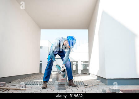 Builder in uniform cutting paving tiles with electric cutter on the construction site with white houses on the background Stock Photo