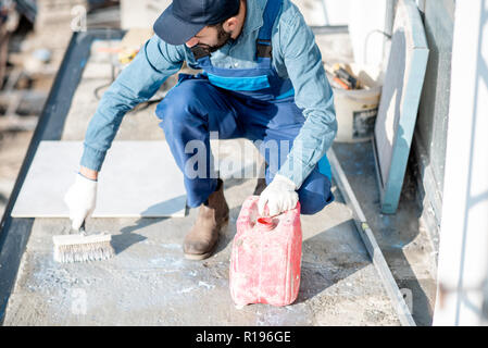 Workman in uniform priming concrete with brush for tiles lying on the balcony Stock Photo