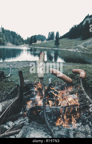 wooden sticks grilling barbecue sausages on fireplace in front of mountain lake Stock Photo