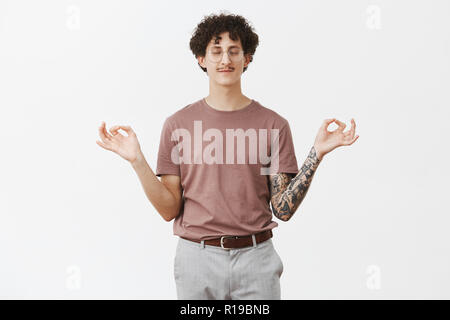 Charming curly-haired guy with funny moustache standing in lotus pose trying find peace and comfort closing eyes and smiling with calm and relaxed exp Stock Photo