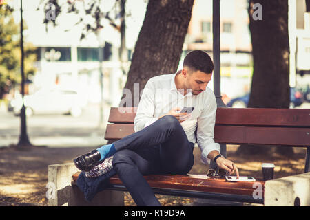 A stylish businessman is sitting on a bench in the park with crossed legs, he is holding his mobile phone and checking something at his tablet Stock Photo