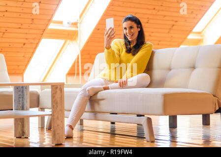 Portrait of a beautiful woman taking a selfie photo her the phone while relaxing on the sofa in a wooden attic loft. Stock Photo